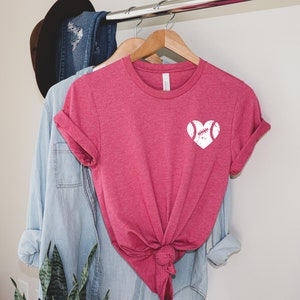 May include: A pink t-shirt with a white baseball heart graphic. The shirt is on a hanger and has a knotted front.