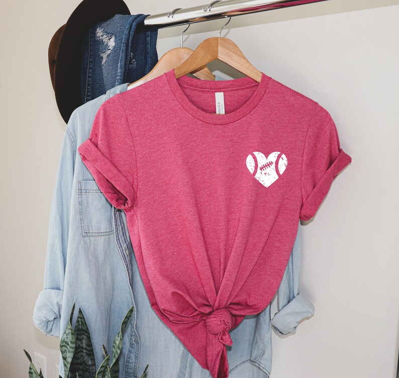May include: A pink t-shirt with a white baseball heart graphic. The shirt is on a hanger and has a knotted front.