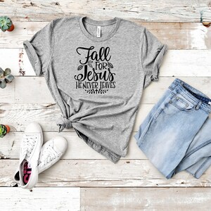 K&ouml;nnte beinhalten: Graues T-Shirt mit dem Text "Fall for Jesus He Never Leaves" in schwarzer Schrift. Das Shirt liegt auf einer wei&szlig;en Holzoberfl&auml;che mit einem Paar wei&szlig;en Sneakers, einer blauen Jeans und kleinen Topfpflanzen.
