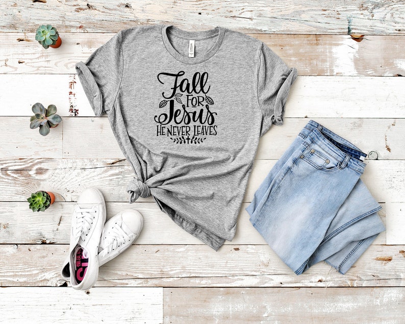 K&ouml;nnte beinhalten: Graues T-Shirt mit dem Text "Fall for Jesus He Never Leaves" in schwarzer Schrift. Das Shirt liegt auf einer wei&szlig;en Holzoberfl&auml;che mit einem Paar wei&szlig;en Sneakers, einer blauen Jeans und kleinen Topfpflanzen.