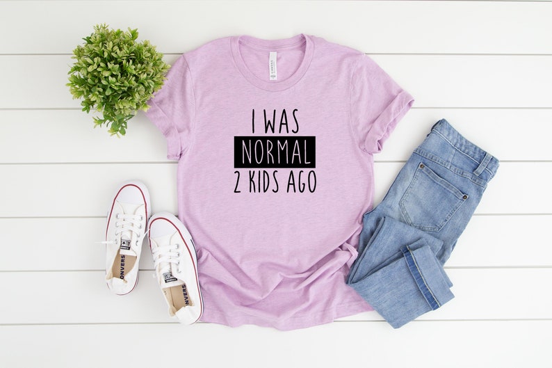 May include: A light purple t-shirt with black text that reads "I was normal 2 kids ago". The shirt is laid flat on a white wooden surface with a pair of white sneakers and blue jeans.