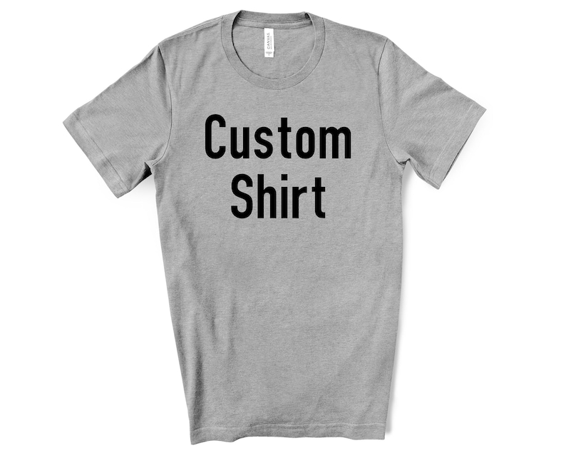 May include: A heather grey short-sleeve t-shirt featuring the black text Custom Shirt printed in the centre of the chest. The garment has a crew neckline and a standard fit, with a white brand label visible inside the collar area.