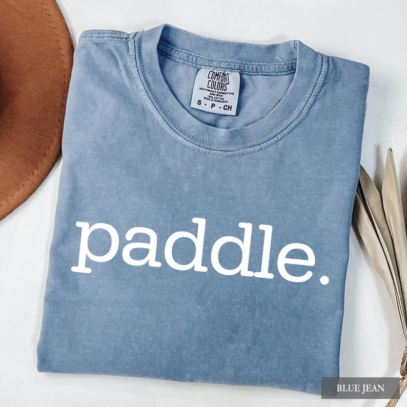 A folded blue jean colored t-shirt with the word paddle printed in white across the chest.