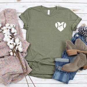 May include: A green t-shirt with a white distressed heart design featuring a football inside. The shirt is displayed on a white wood background with a pair of jeans, a sweater, and a pine cone.