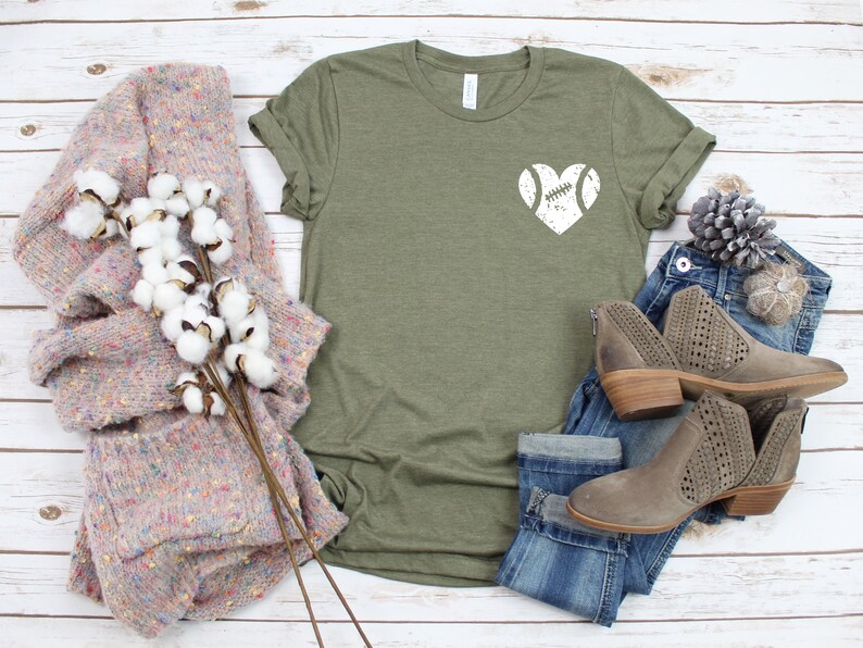 May include: A green t-shirt with a white distressed heart design featuring a football inside. The shirt is displayed on a white wood background with a pair of jeans, a sweater, and a pine cone.