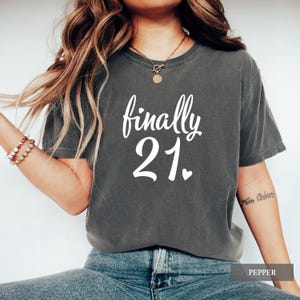 Peut inclure: Un t-shirt gris foncé avec l'inscription blanche "finally 21", avec un petit cœur sous le chiffre 21. Le t-shirt est de style décontracté, adapté à une fête d'anniversaire. Le mot "PEPPER" est dans un rectangle en bas.