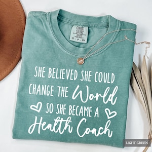 May include: Light green Comfort Colors t-shirt with the text "SHE BELIEVED SHE COULD CHANGE THE WORLD SO SHE BECAME A Health Coach" in white. A gold necklace with a pendant is on the shirt. The shirt is labeled "LIGHT GREEN".
