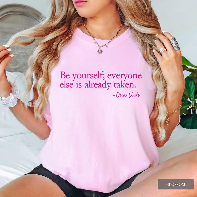 A person wears a light pink t-shirt printed with the quote "Be yourself; everyone else is already taken. - Oscar Wilde" in dark pink lettering.