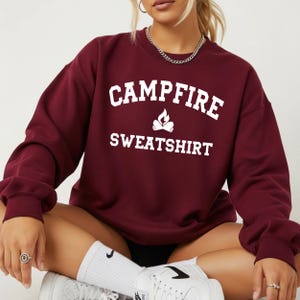 A person wearing a maroon crewneck sweatshirt with "CAMPFIRE SWEATSHIRT" in white text and a small campfire icon, sits with crossed legs.