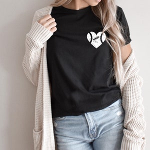 May include: A black t-shirt with a white graphic of a baseball in a heart shape. The shirt is being worn by a person with long blonde hair and a cream cardigan.