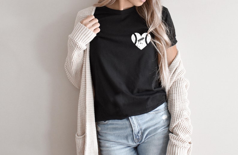 May include: A black t-shirt with a white graphic of a baseball in a heart shape. The shirt is being worn by a person with long blonde hair and a cream cardigan.