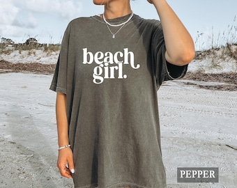 Può includere: Una maglietta verde oliva scuro con la scritta "beach girl" in bianco. La maglietta ha una vestibilità ampia e sembra essere realizzata in un materiale morbido. La parola "pepper" è in un riquadro bianco in basso a destra.