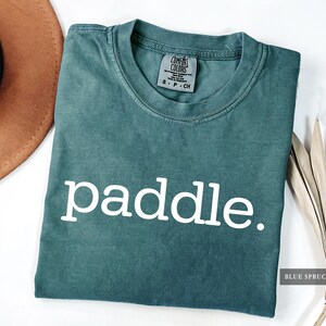 A folded blue spruce colored t-shirt with the word paddle printed in white across the chest.