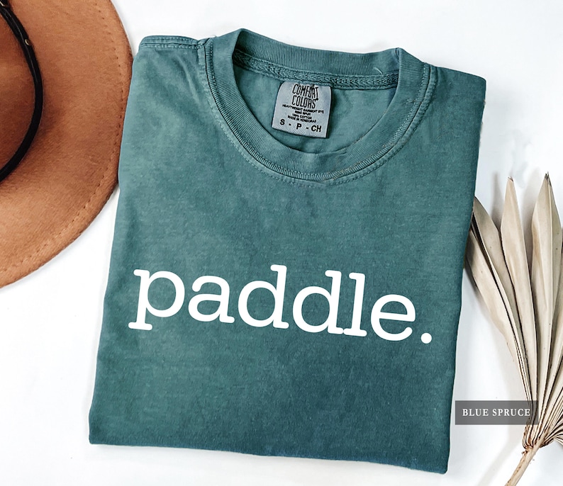 A folded blue spruce colored t-shirt with the word paddle printed in white across the chest.