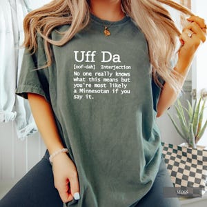 May include: A moss green t-shirt with the text "Uff Da" in white, along with a definition. The shirt is a casual style, suitable for everyday wear. The text explains the meaning of the phrase.