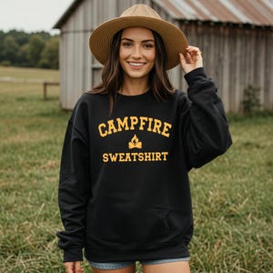 A woman wearing a straw hat and a black crewneck sweatshirt with "CAMPFIRE SWEATSHIRT" in yellow text and a small campfire icon, stands in a field.