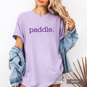 A person wearing an orchid colored t-shirt with the word paddle printed in purple on the chest and a denim jacket draped over one arm.