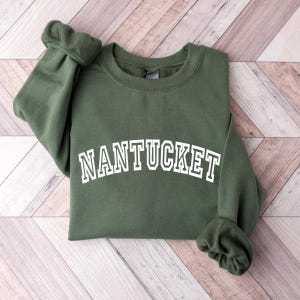 Puede incluir: Una sudadera de cuello redondo en color verde oliva con la palabra NANTUCKET impresa en blanco con una tipograf&iacute;a arqueada de estilo universitario en el pecho. La prenda tiene mangas largas con pu&ntilde;os acanalados y un cuello acanalado sencillo.