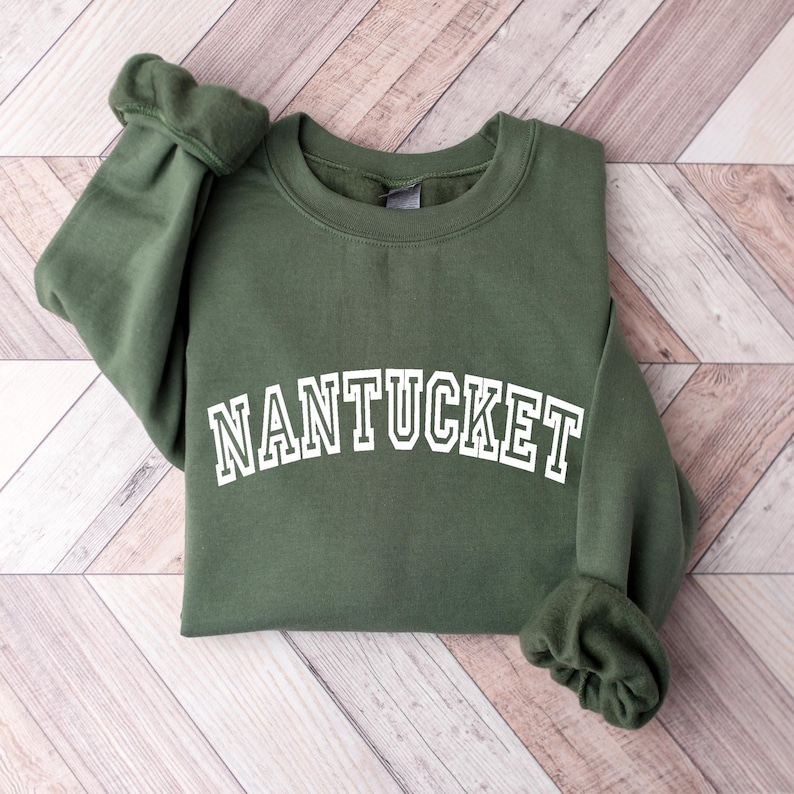 Puede incluir: Una sudadera de cuello redondo en color verde oliva con la palabra NANTUCKET impresa en blanco con una tipograf&iacute;a arqueada de estilo universitario en el pecho. La prenda tiene mangas largas con pu&ntilde;os acanalados y un cuello acanalado sencillo.