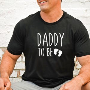 May include: Black crew neck t-shirt with the text "DADDY TO BE" in white, along with two small white footprints. The shirt is made of a soft material.