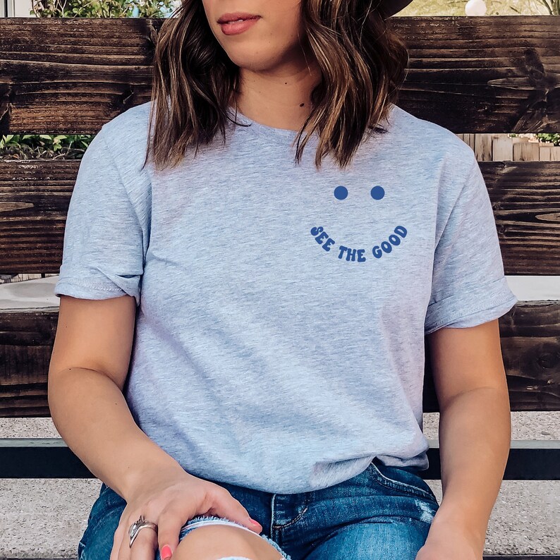 K&ouml;nnte beinhalten: Ein hellgraues T-Shirt mit einem blauen Smiley und dem Text "See the Good" auf der Vorderseite.