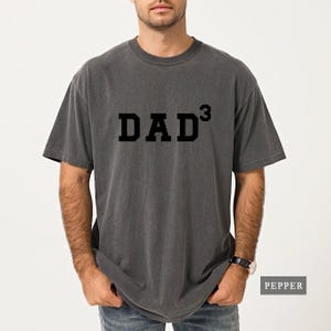 May include: Dark gray t-shirt with the word "DAD³" printed in black block letters. The shirt has short sleeves and a crew neck. The word "PEPPER" is printed in a white rectangle in the lower right corner.