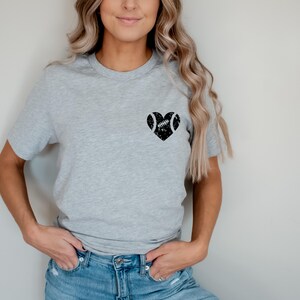 May include: A gray t-shirt with a black heart design featuring a football inside. The heart is centered on the chest of the shirt.