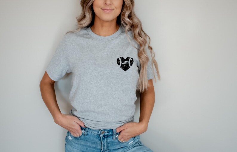May include: A gray t-shirt with a black heart design featuring a football inside. The heart is centered on the chest of the shirt.