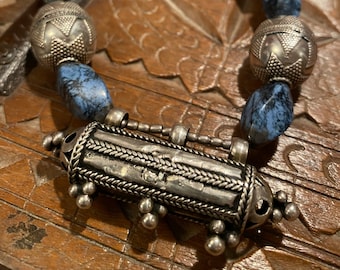 May include: A silver necklace with a long, cylindrical pendant and blue stone beads. The pendant has intricate designs and is attached to the necklace with a silver chain.