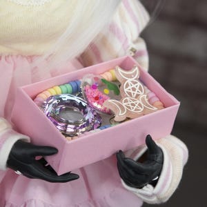 Könnte beinhalten: Eine Puppe in einem hellgelben Oberteil mit Spitzenausschnitt und einem rosa Rock, die eine rosa Schachtel mit bunten Accessoires hält. Die Schachtel enthält ein regenbogenfarbenes Perlenarmband, einen herzförmigen Anhänger und andere dekorative Gegenstände.