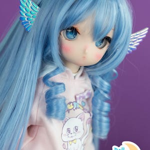 Può includere: Una bambola con i capelli blu che indossa una felpa rosa con un'immagine di un gatto bianco e il testo "Doll Moon". La bambola ha gli occhi blu e indossa delle ali iridescenti su ogni orecchio.