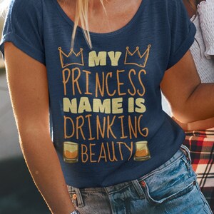 Op de afbeelding: Twee vriendinnen lachen en houden drankjes vast. Een van hen draagt een blauw shirt met de tekst "My Princess Name is Drinking Beauty" erop gedrukt.