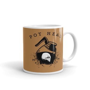 May include: A white ceramic coffee mug with a brown background. The mug has a black and white illustration of a coffee pot with a skull inside. The text "POT HEAL" is printed on the mug.