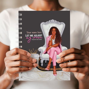 May include: A spiral-bound notebook with a black cover featuring an illustration of a woman in a pink suit seated on a throne. The text "LET ME ADJUST my crown" is displayed. The notebook is held by hands.
