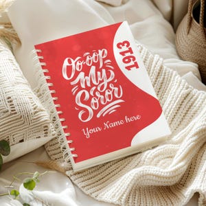 May include: A red spiral-bound notebook with the text "Oo-oop My Soror" in white script. The notebook also features the year "1913" and the words "Your Name here". The notebook is lying on a cream-colored knitted blanket.