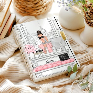 May include: A white spiral bound notebook with a black and white illustration of a woman in a pink jacket and sunglasses sitting at a desk in front of a city skyline. The notebook has the text "Name" and "Good Vibes Only" on the cover.