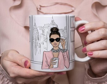 May include: White ceramic mug with a pink and black illustration of a woman wearing sunglasses and a pink jacket. The woman is holding a coffee cup and is standing in front of a city skyline. The mug has a black handle.