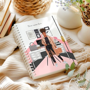 May include: Spiral-bound notebook featuring an illustration of a woman in a stylish office setting. The cover includes the text "Your name here" and a depiction of a desk with a computer, purse, and coffee cup. The design incorporates pink and black geometric shapes.