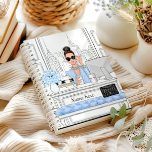 May include: A white spiral bound notebook with a black and white illustration of a woman in a blue outfit sitting at a desk with a city skyline in the background. The notebook has a blue and white patterned bench with the text "Name here" on it. The notebook also has a black and white book with the text "History of Fashion" on it.