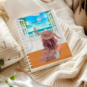 May include: A spiral-bound notebook with a beach scene illustration. The cover features a woman looking out a window at a tropical ocean view. The text "Name here" is written on the cover. The notebook is resting on a cream-colored blanket.