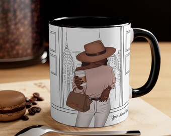 May include: A white ceramic mug with a black rim and handle. The mug features a black and white illustration of a woman wearing a brown hat and a pink sweater, holding a coffee cup. The background is a city skyline. The text "Your Name Here" is printed on the bottom right corner of the mug.