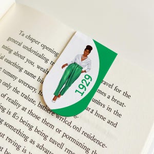 May include: A bookmark featuring an illustration of a person in a white blouse and green pants. The number "1929" is printed in green. The bookmark is placed on the pages of a book.