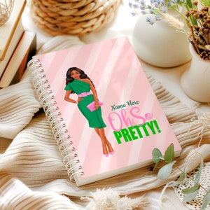 May include: Spiral-bound notebook with a pink and white striped cover. The cover features an illustration of a woman in a green dress and the text "Name Here Oh So Pretty!"