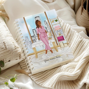May include: A spiral-bound planner with a cover illustration of a woman in a pink jumpsuit standing in an office with a city view. The cover has the text "Your name here" and a marble design.