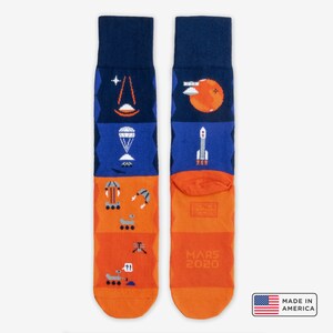 May include: A pair of orange, blue, and white socks with a space exploration theme. The socks feature images of a rocket, a rover, a parachute, and the text "Mars 2020".