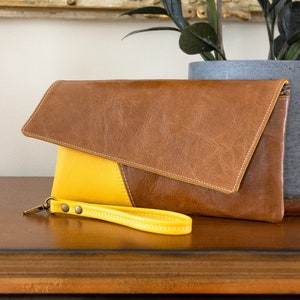 May include: A brown leather clutch with a yellow leather flap and a yellow leather wrist strap. The clutch is on a wooden surface.