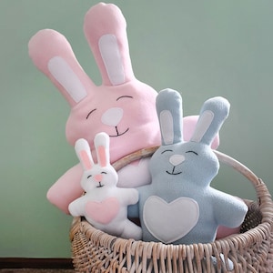 Op de afbeelding: Drie pluche konijnen speelgoed in een rieten mand. Het grootste konijn is roze met witte oren en een wit hart op zijn borst. Het middelste konijn is blauw met witte oren en een wit hart op zijn borst. Het kleinste konijn is wit met roze oren en een roze hart op zijn borst.