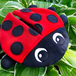 May include: A plush ladybug toy with black spots and white eyes. The ladybug is red with black spots and has a black head with white eyes. The toy is sitting on a bed of green leaves.