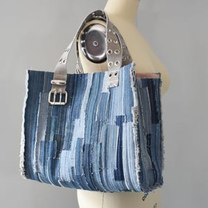 May include: A patchwork denim tote bag in shades of blue. The bag has silver-tone buckle and rivet details on the straps. The raw edges of the denim give it a rustic, handcrafted look. The bag is approximately 35 cm wide and 30 cm tall.