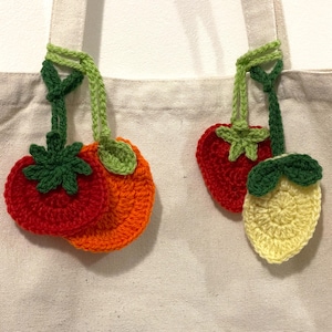 Puede incluir: Primer plano de un bolso tote beige con abalorios de frutas de ganchillo. Los abalorios incluyen una pila de tomates rojos y naranjas, una fresa roja y un limón amarillo. Cada abalorio tiene tallos y hojas verdes.